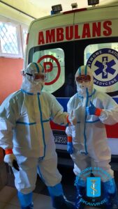 DWÓCH RATOWNIKÓW W STROJACH OCHRONY BIOLOGICZNEJ STOI W GARAŻU PRZY KARETCE